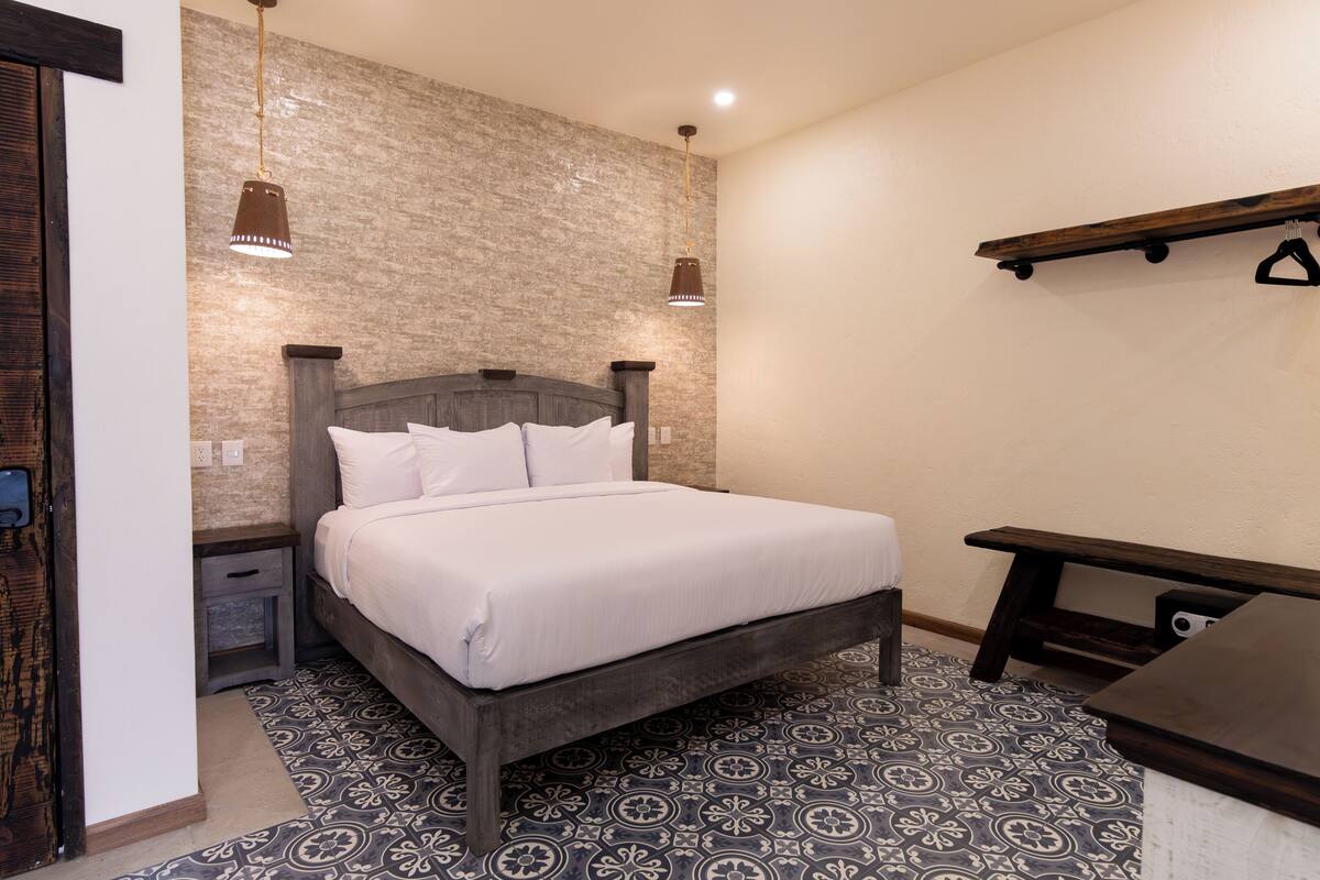 A bedroom with a bed, bedside tables, a hanging light fixture, a wooden bench, and patterned floor tiles.