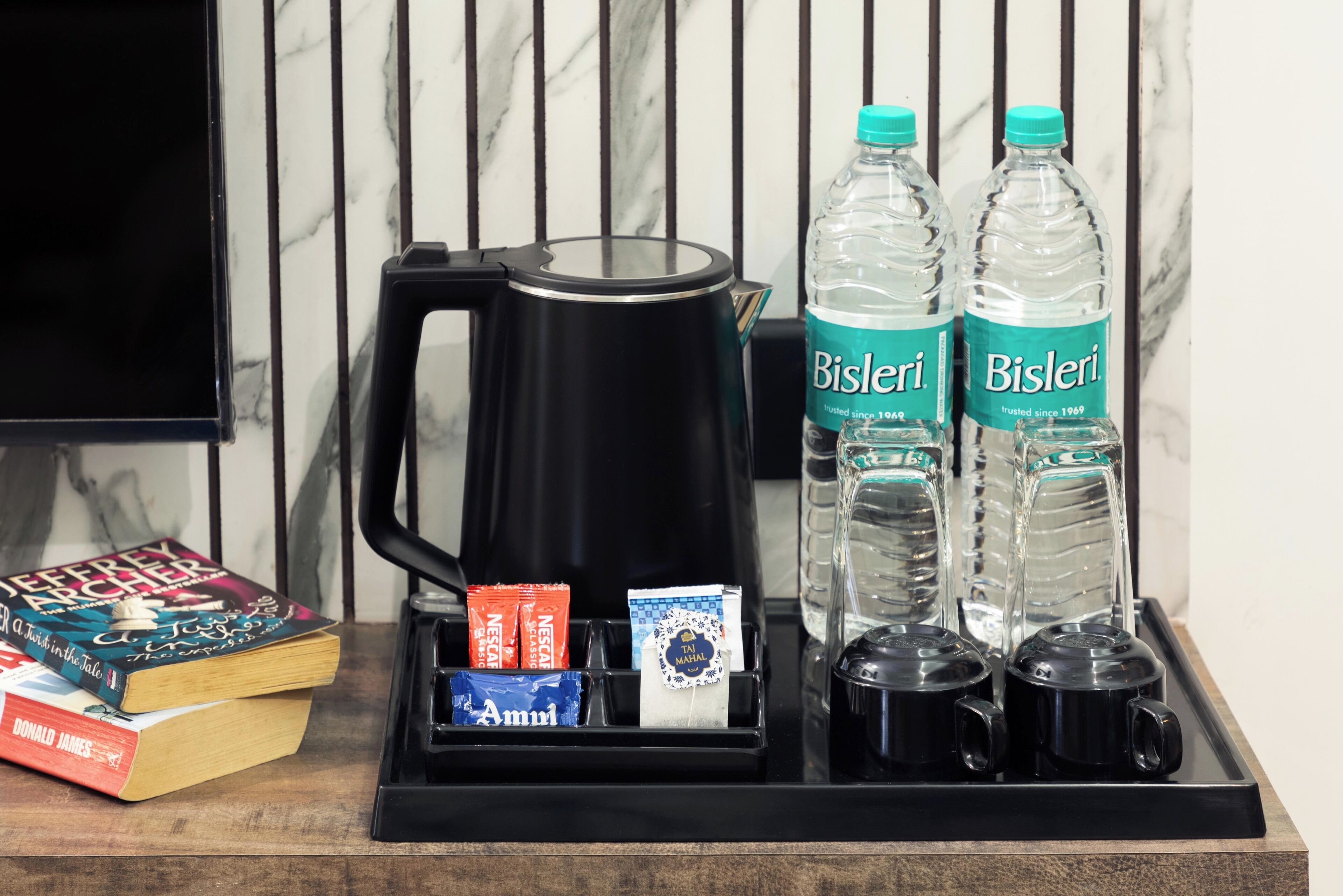 A tray with a black coffee maker, two Bisleri water bottles, a packet of Amul tea, and a book titled 'Jeffery Archer'.