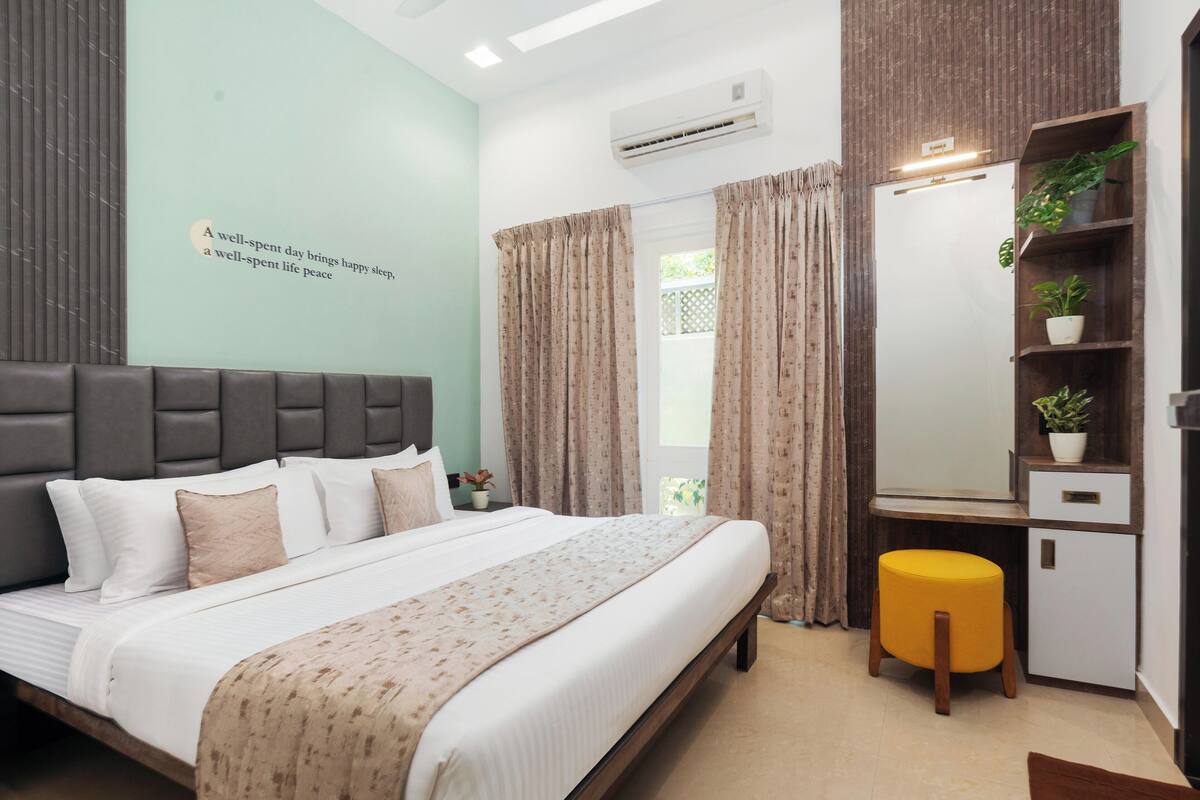 A modern bedroom with a large bed, a bedside table with a lamp, a small stool, a wardrobe, and a wall-mounted air conditioner.