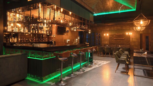 A bar area with a wooden bar counter, multiple stools, and a variety of bottles and glasses. - KAIRA PARK (Chennai)