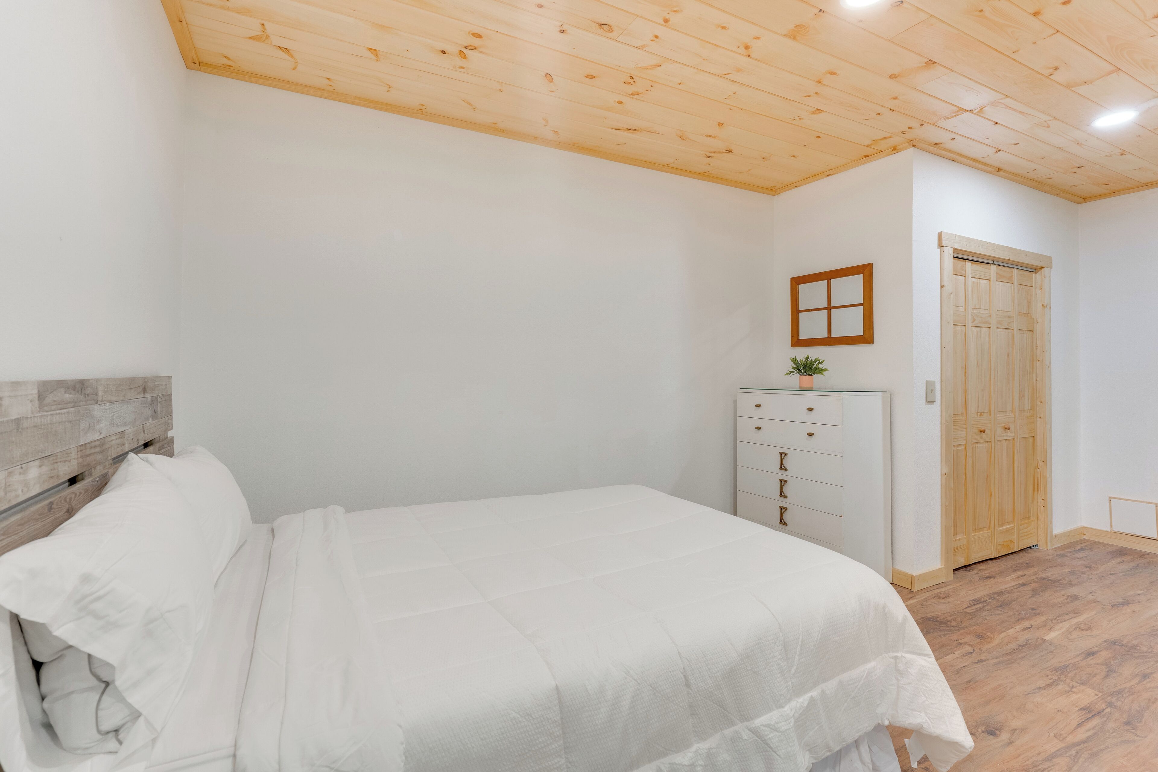 Camera da letto con soffitto in legno, un letto con biancheria bianca, una cassettiera, una porta in legno e una piccola pianta in vaso.