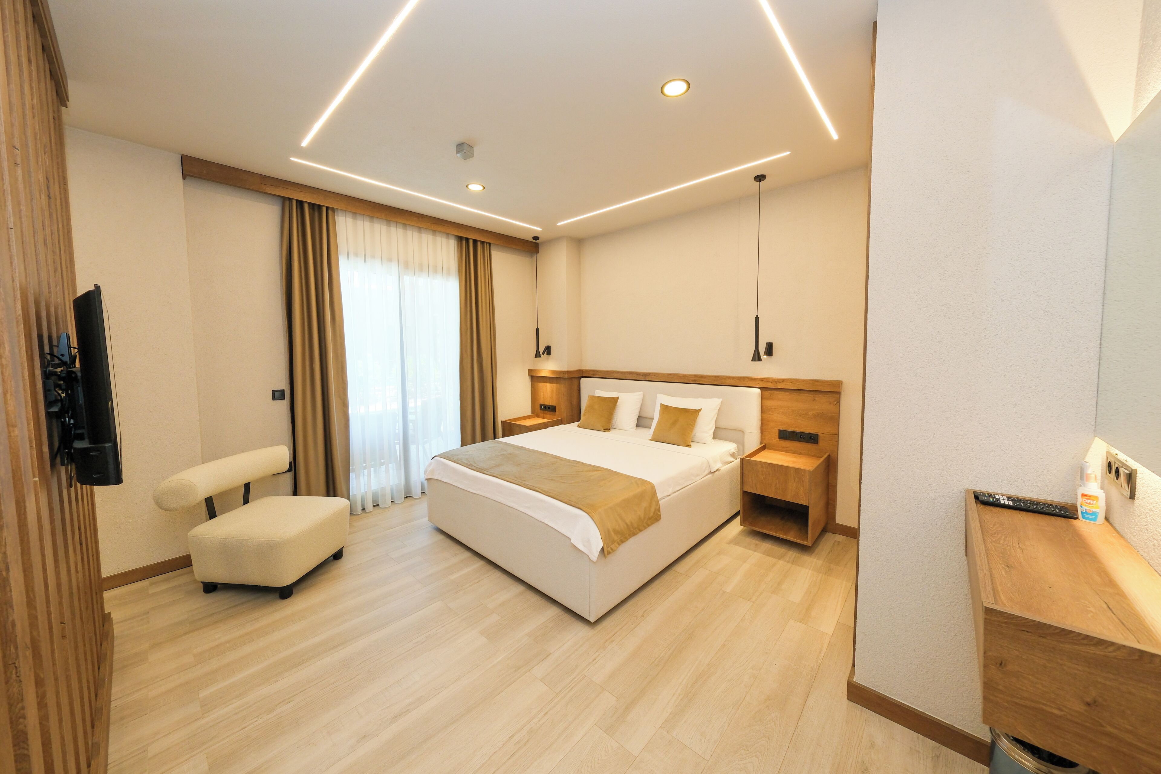 A modern hotel room with a large bed, bedside tables, a flat-screen TV, a sofa, and a wooden bench.