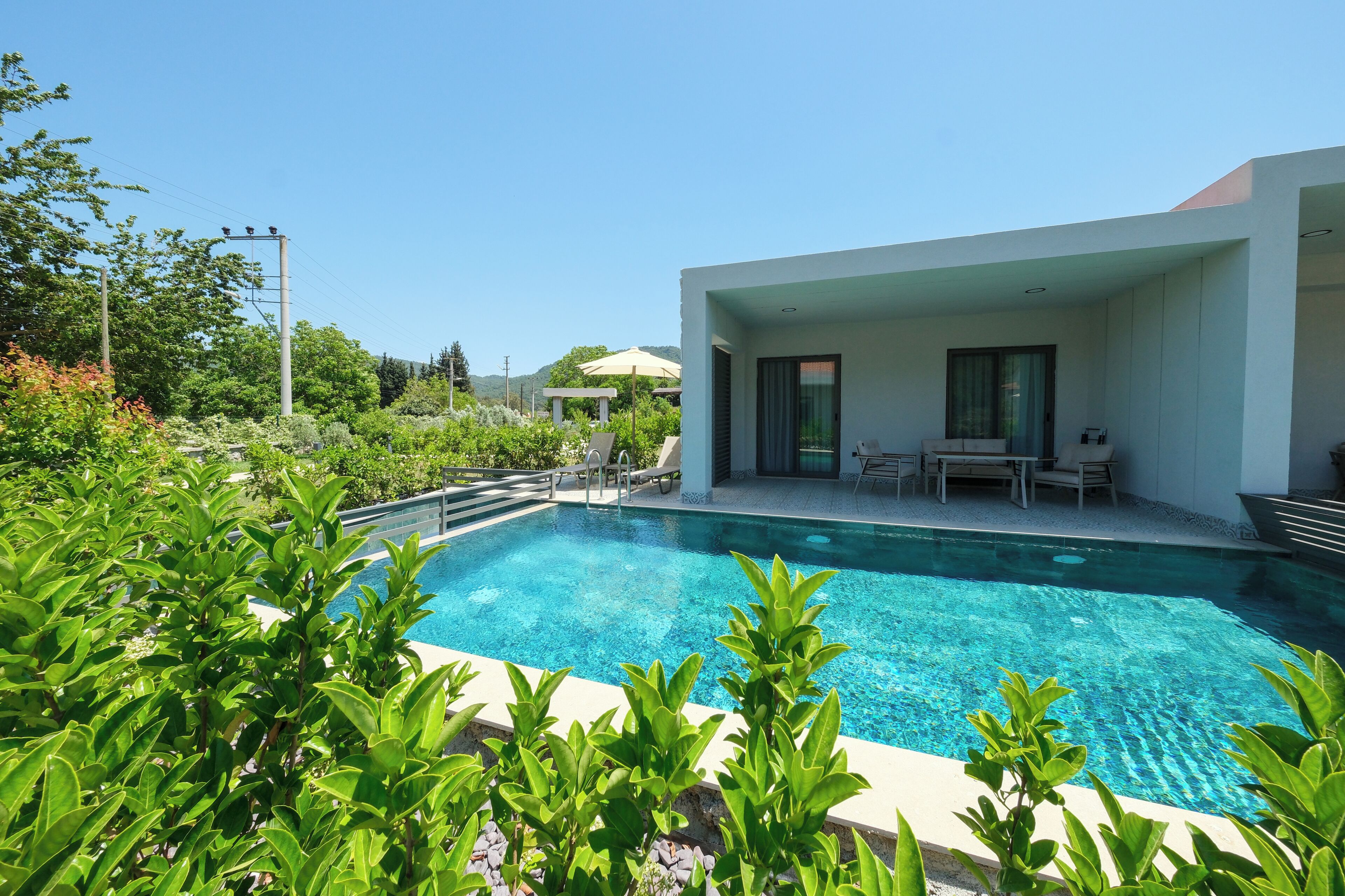 Un área moderna junto a la piscina con agua cristalina, mobiliario exterior y abundante vegetación.