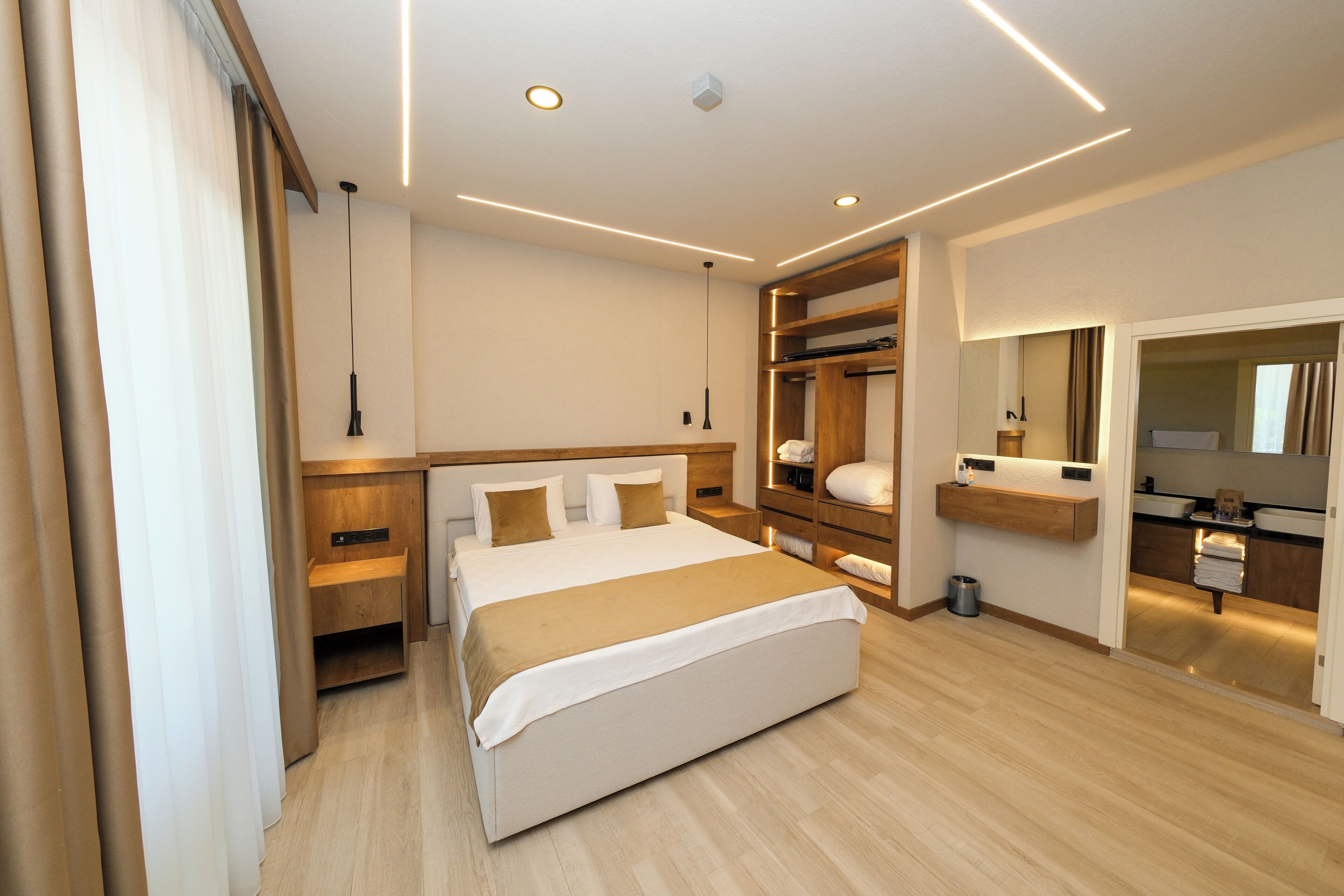 Une chambre d’hôtel moderne avec un grand lit, une tête de lit en bois, des tables de chevet, une armoire intégrée, une coiffeuse avec un miroir et une petite poubelle.