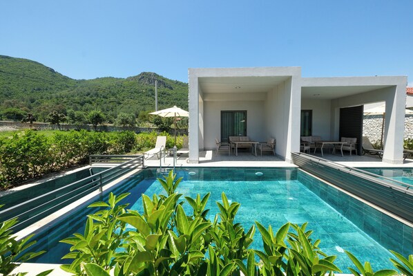 A modern pool area with clear blue water, surrounded by lush greenery and a mountainous backdrop. - Casa Dila (Marmaris)