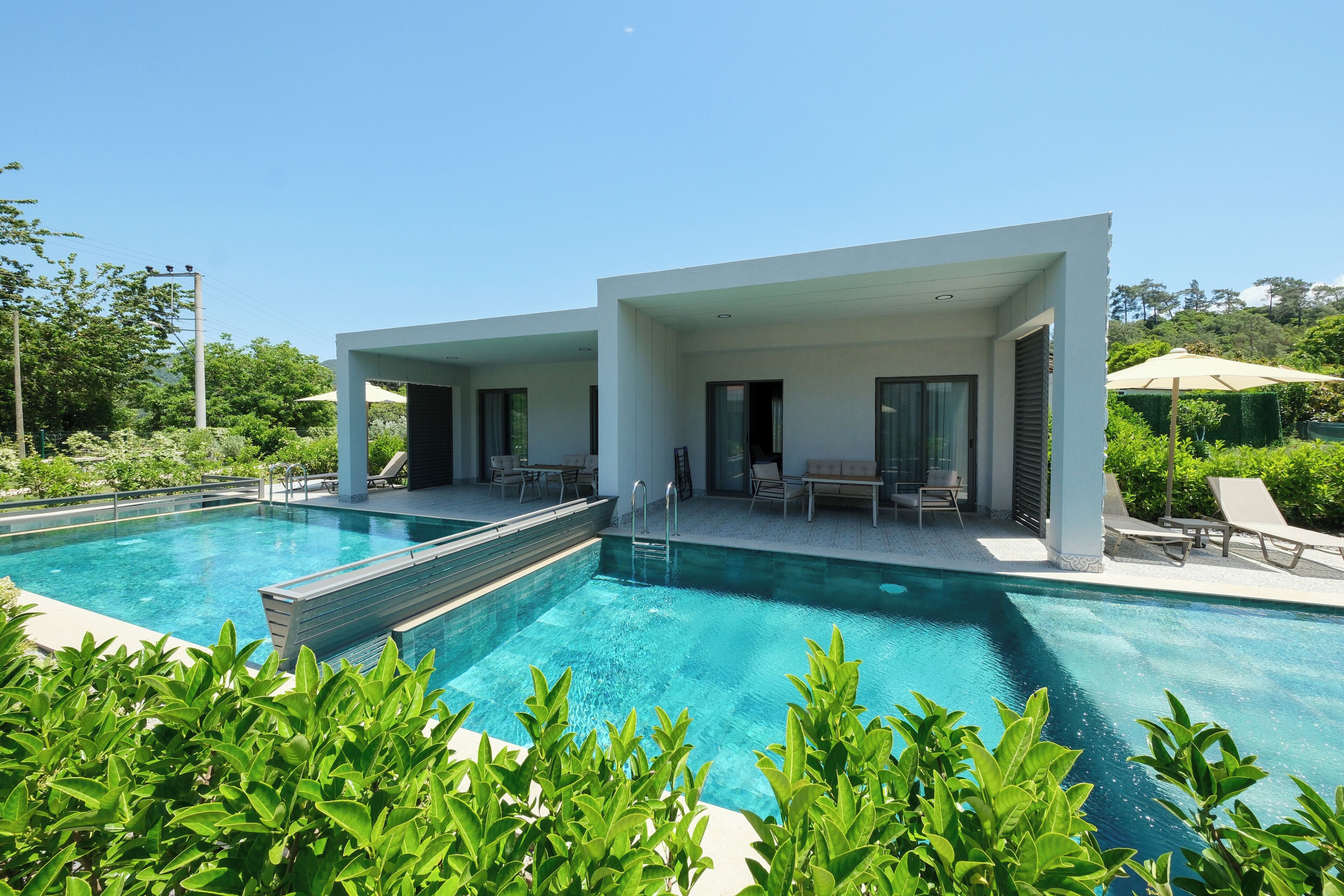 Un área de piscina moderna con agua cristalina, rodeada de exuberante vegetación y un edificio blanco con grandes ventanales.
