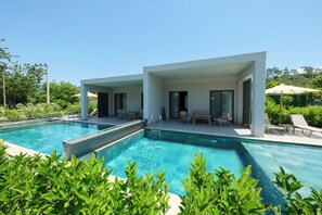 Un espace piscine moderne avec une eau cristalline, entouré d’une végétation luxuriante et d’un bâtiment blanc doté de grandes fenêtres.