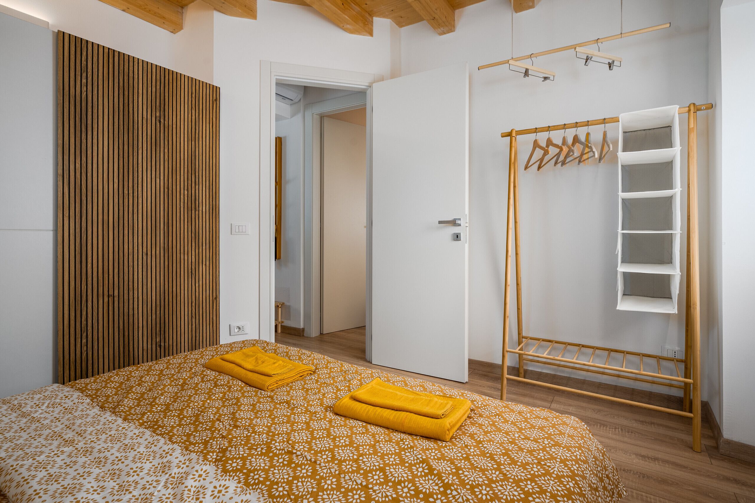 Une chambre avec un lit, un mur recouvert de lambris en bois, une armoire, un étendoir et une porte donnant sur une autre pièce.