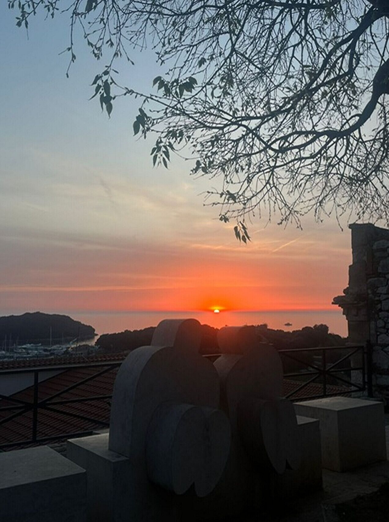 Vista del atardecer desde un balcón con vistas a una masa de agua, con un edificio de piedra y un árbol en primer plano.