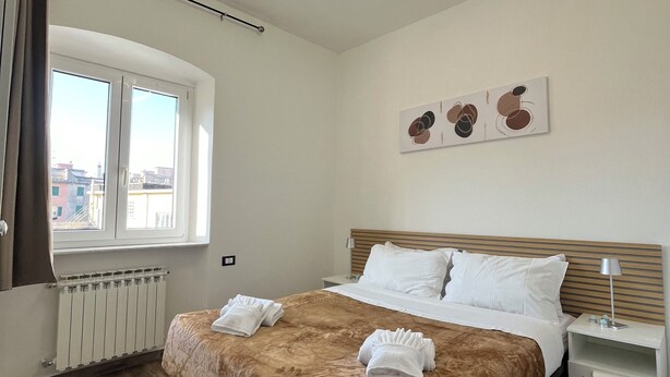 Hotellihuone, jossa on suuri sänky, ikkuna verhoilla, patteri, yöpöytä lampun kanssa ja seinällä taideteos.