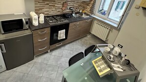 Kompakti keittiö moderneilla laitteilla, ruokailutila lasipöydällä sekä ikkuna, josta avautuu näkymä rakennuksiin.