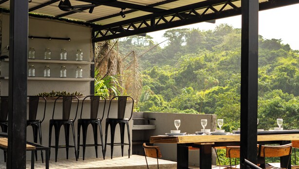 A covered outdoor dining area with metal chairs and a table set for two, overlooking a lush green forest. - Casa Xué: Soul of Tayrona (La Poza)