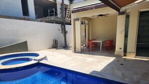Una piscina con diseño circular, un espacio de patio cubierto con mobiliario exterior y un edificio de estilo arquitectónico moderno.