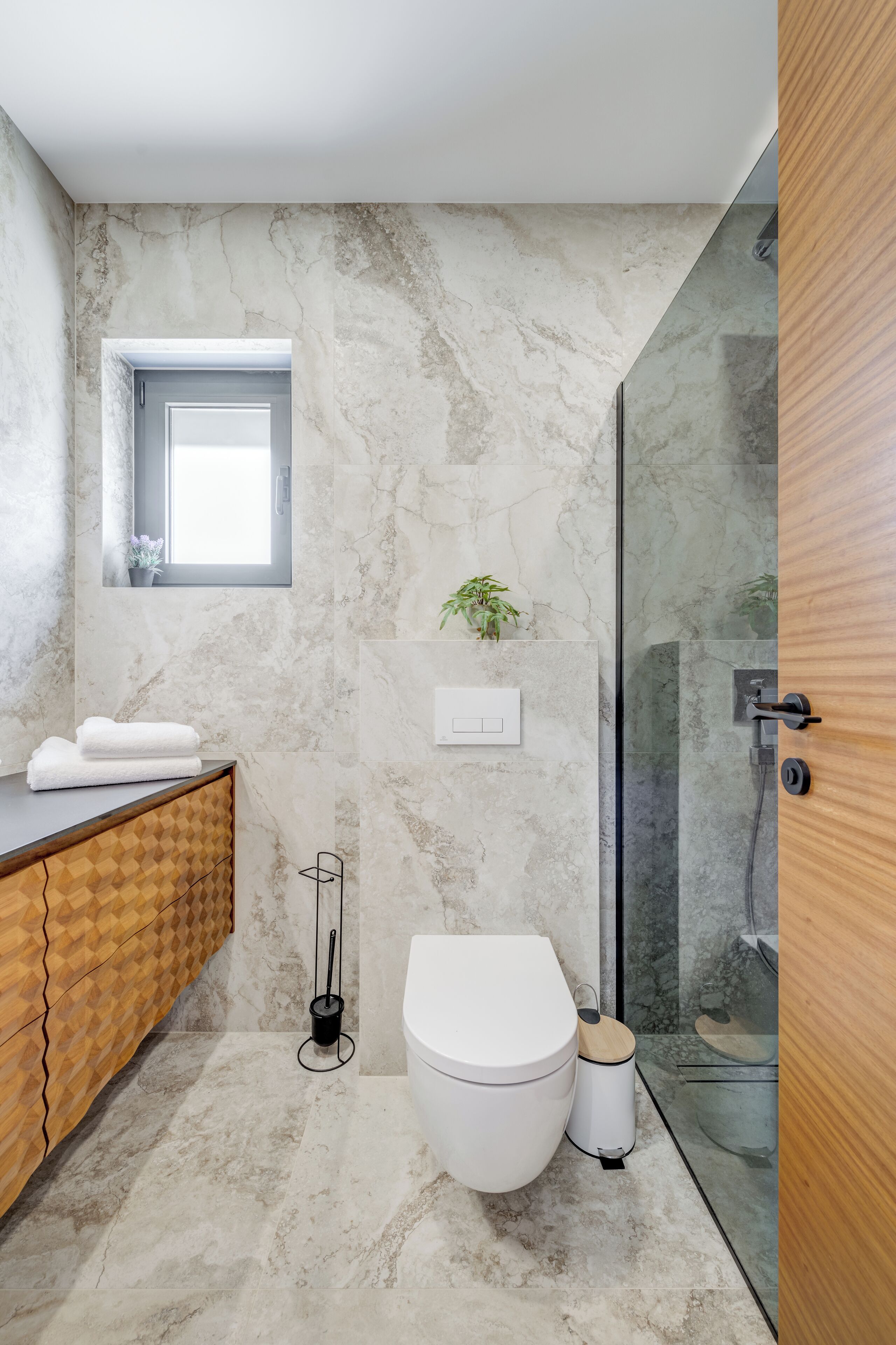 A modern bathroom with a marble wall, a wooden vanity, a toilet, a glass shower enclosure, and a small window.