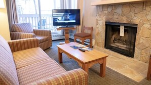 A living room with a stone fireplace, a television, and patterned furniture. - 2 Bedroom Condo @ Banff Rocky Mountain Resort - June 14 to June 21, 2026 (Banff)