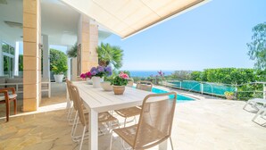 An outdoor patio with a white table and wicker chairs, overlooking a swimming pool and the sea.