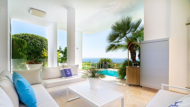 A modern outdoor living area with a white sofa, a coffee table, and a view of a swimming pool and ocean.