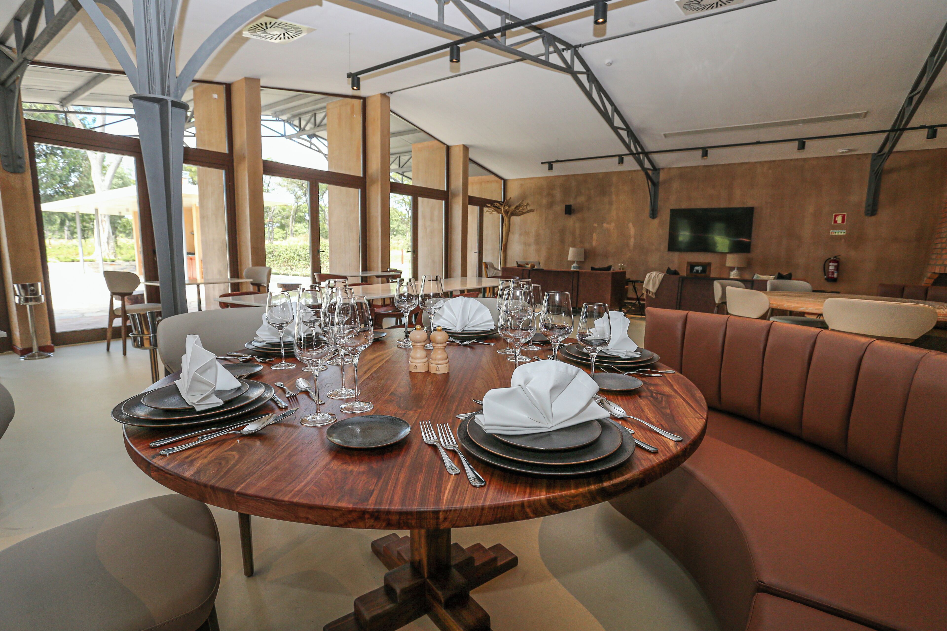 A dining area with a round table set for a meal, surrounded by cushioned seating and large windows.