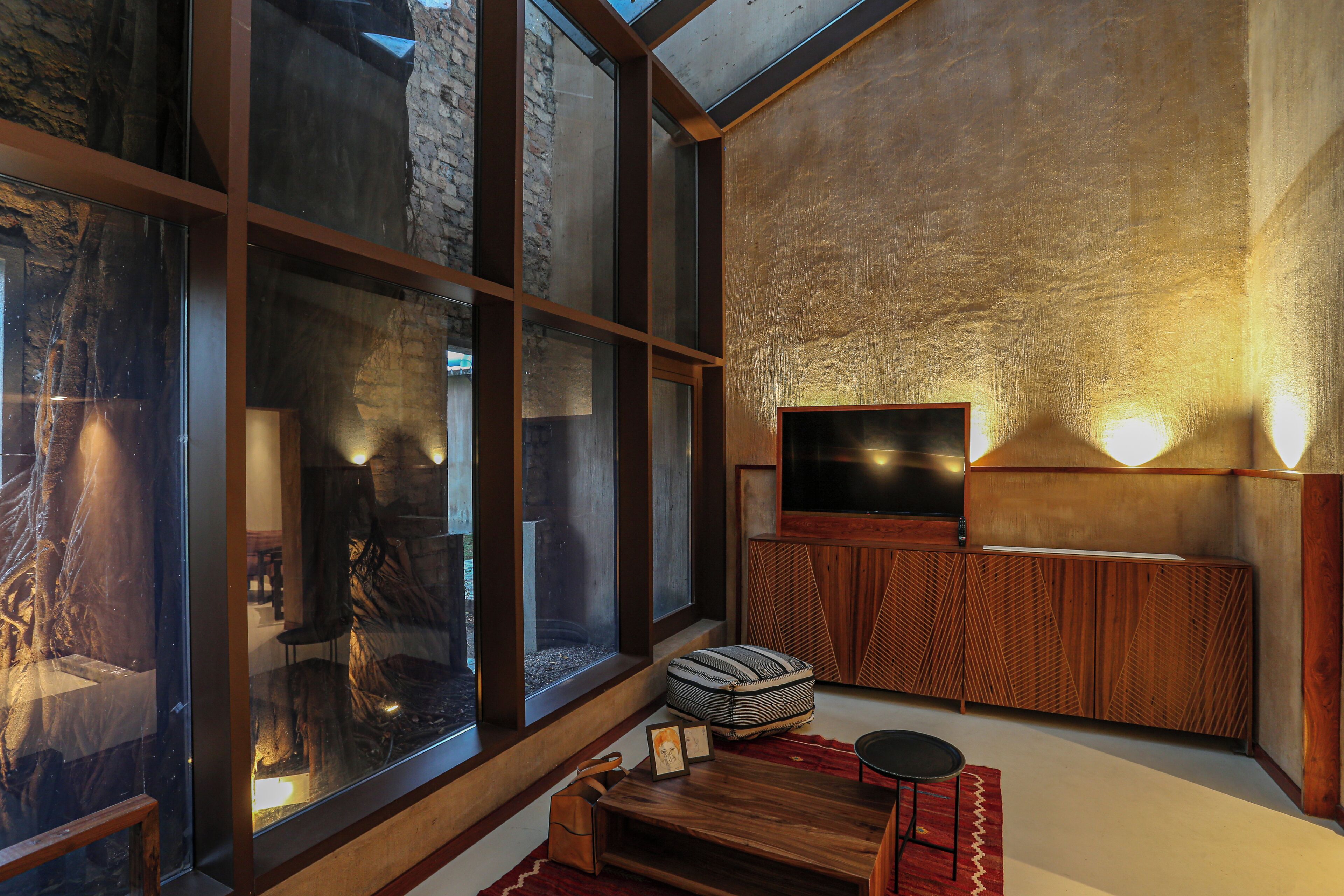 Un intérieur moderne avec de grandes fenêtres, un banc en bois, un téléviseur et un mur orné d’un motif.