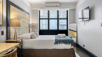 Une chambre d’hôtel avec un grand lit, un bureau, une chaise, une télévision et une fenêtre avec des stores.