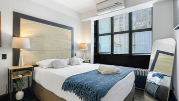 A modern hotel room with a large bed, bedside lamps, a window with blinds, a mirror, and a small seating area.