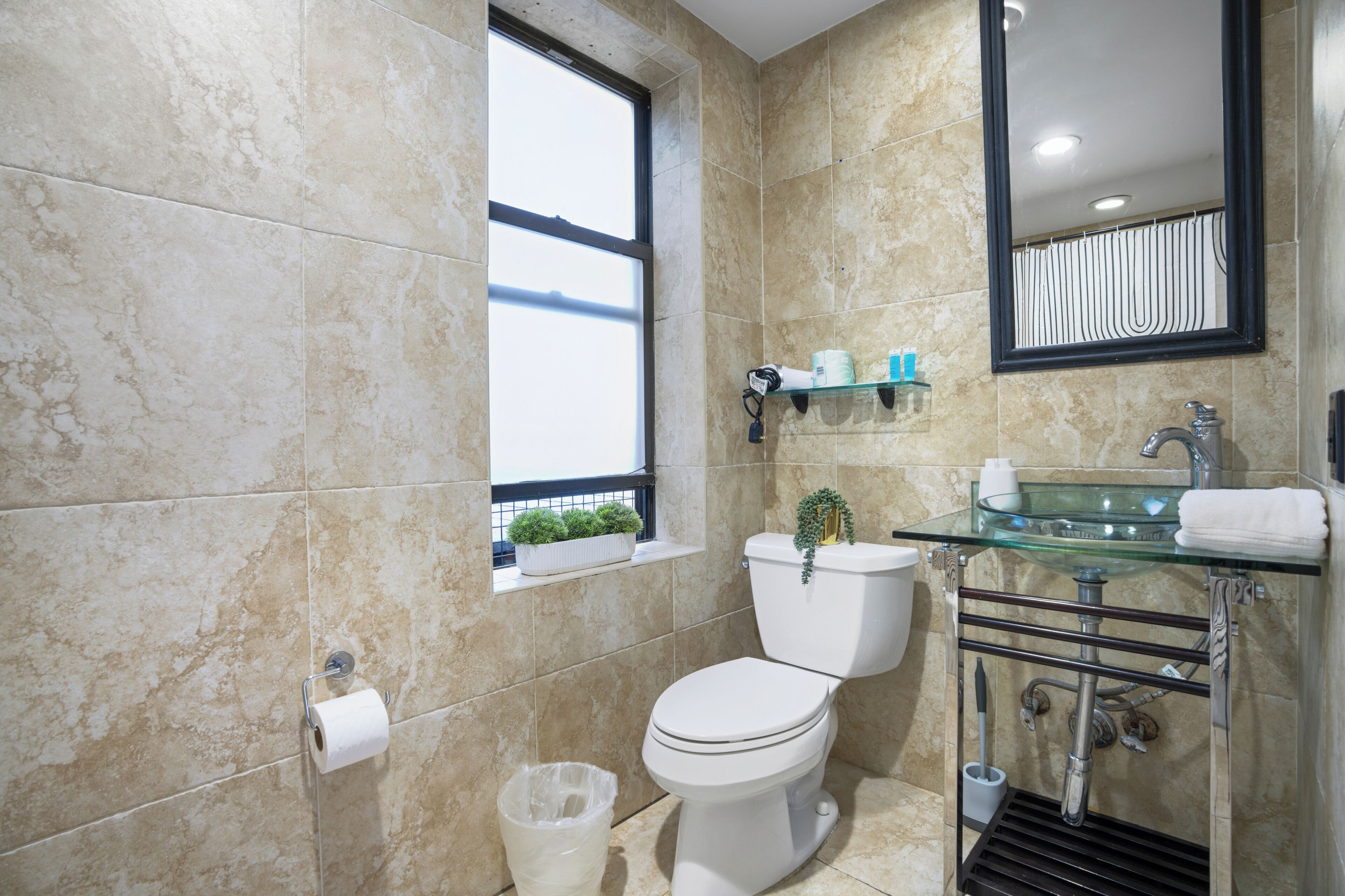 Un baño moderno con inodoro, lavabo de vidrio y una ventana con planta.
