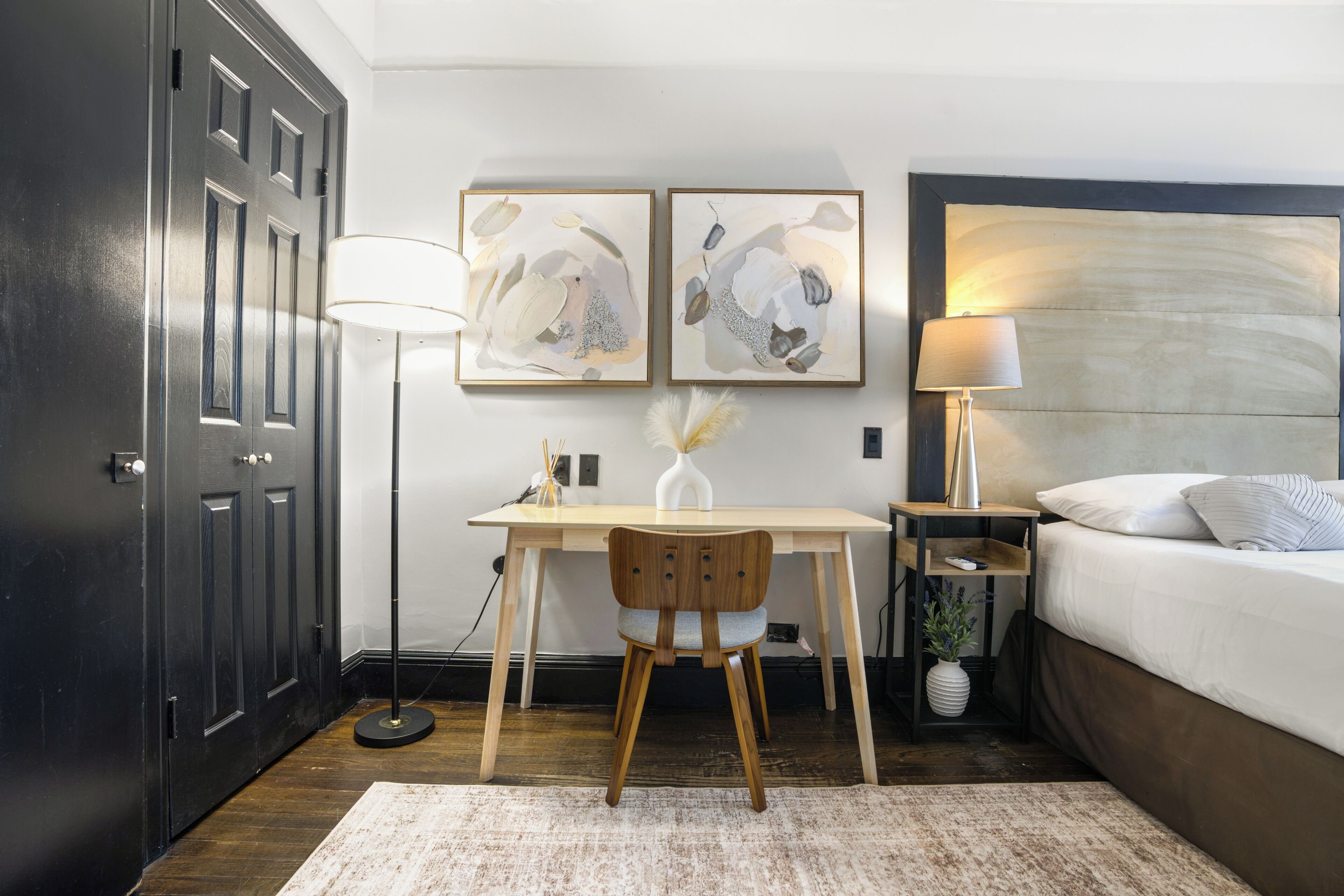 A hotel room with a bed, desk, chair, lamp, and artwork on the wall.
