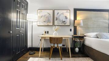A hotel room with a bed, desk, chair, lamp, and artwork on the wall.