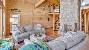 L’intérieur d’un chalet confortable, avec des murs en rondins, une cheminée en pierre, un canapé d’angle gris et un plafond en bois.