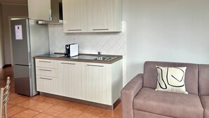 Eine kleine Küche mit Kühlschrank, Toaster und Spüle. Im angrenzenden Wohnbereich befindet sich ein graues Sofa mit einem Kissen.