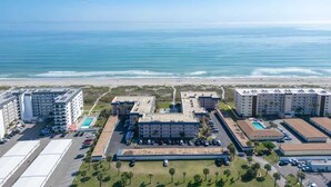 An aerial view of a hotel complex with multiple buildings, a parking lot, and a beach in the background.