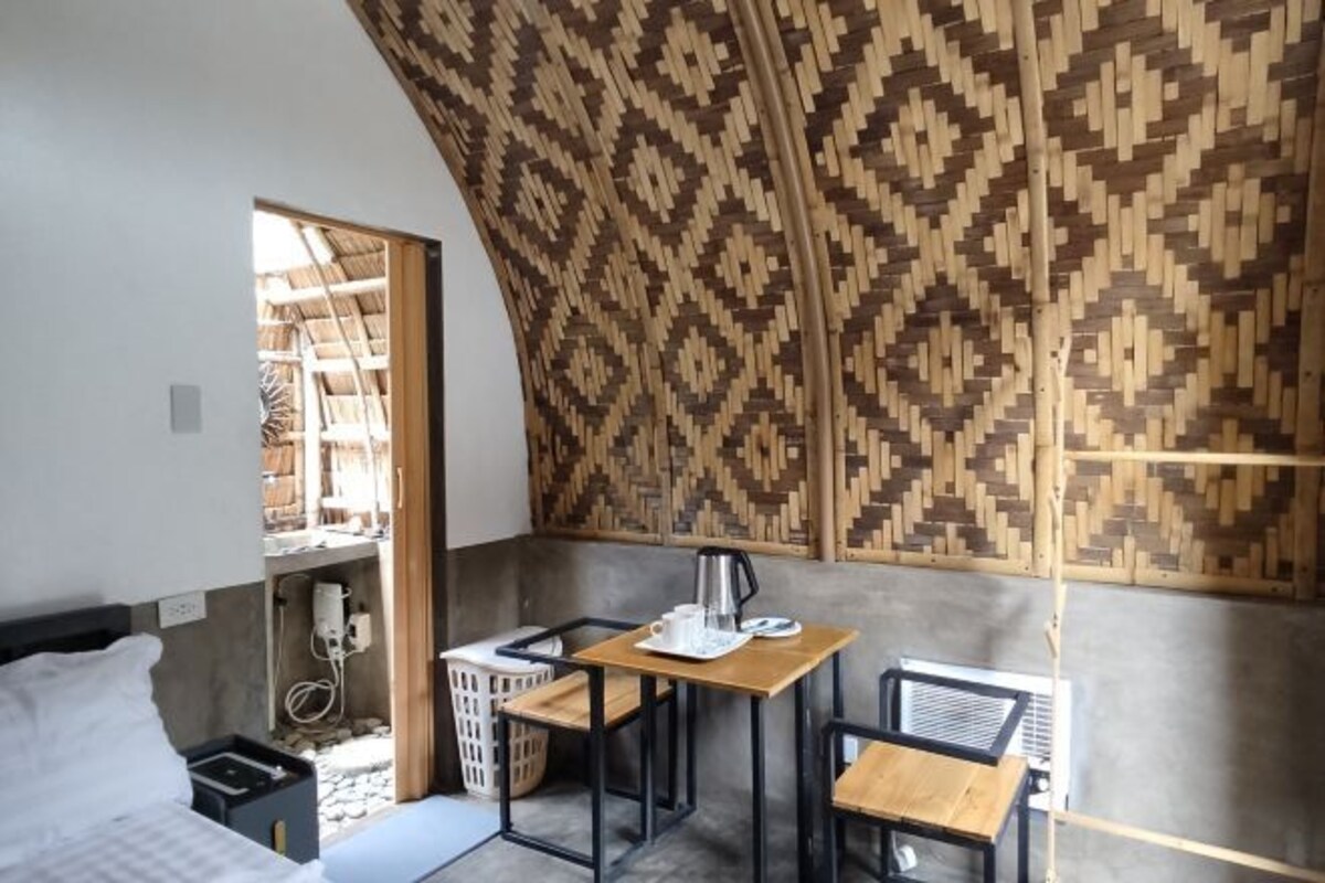 Una habitación compacta con cama, una mesita con hervidor y una silla. Se aprecia una pared con diseño y una ventana con vista a un espacio exterior.