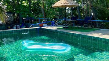 Piscina cristalina con sombrilla de paja, rodeada de exuberante follaje y un suelo de baldosas.