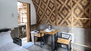 Una habitación con techo alto tejido, una cama con ropa blanca, una mesita con hervidor y una silla de madera.