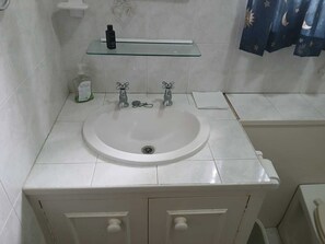 A bathroom with a white sink, two faucets, a glass shelf above the sink, and a white cabinet below.