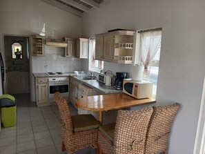 A compact kitchen with white cabinets, a wooden dining table, wicker chairs, and a trash bin.