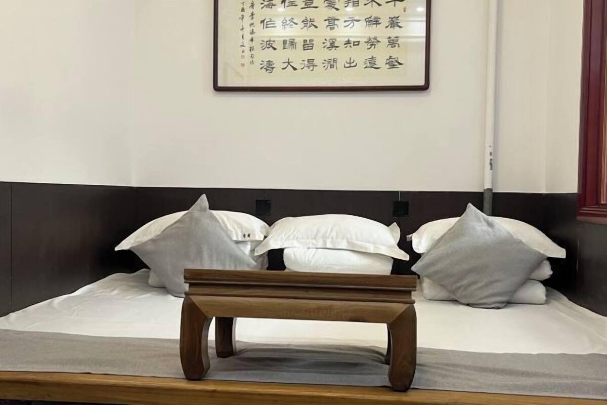 Una cama bien hecha con ropa de cama y almohadas blancas, una mesita de noche de madera y una obra de caligrafía enmarcada en la pared.