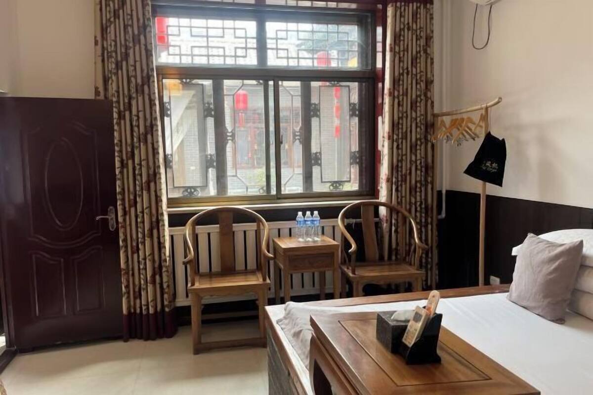 Una habitación de hotel con una cama, dos sillas de madera, una mesita, una ventana con cortinas, una puerta y un teléfono en la pared.