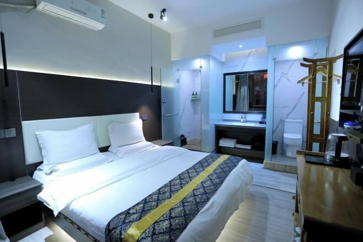 Una habitación de hotel con una cama grande, una mesita de noche, un espejo, un aparador de madera y un baño con lavamanos e inodoro.
