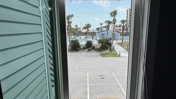 Une vue à travers une fenêtre donnant sur un parking avec une piscine et un petit bâtiment, le tout entouré de palmiers et d’autres immeubles.