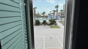 A view through a window of a parking lot with a pool and a small building, surrounded by palm trees and other buildings.