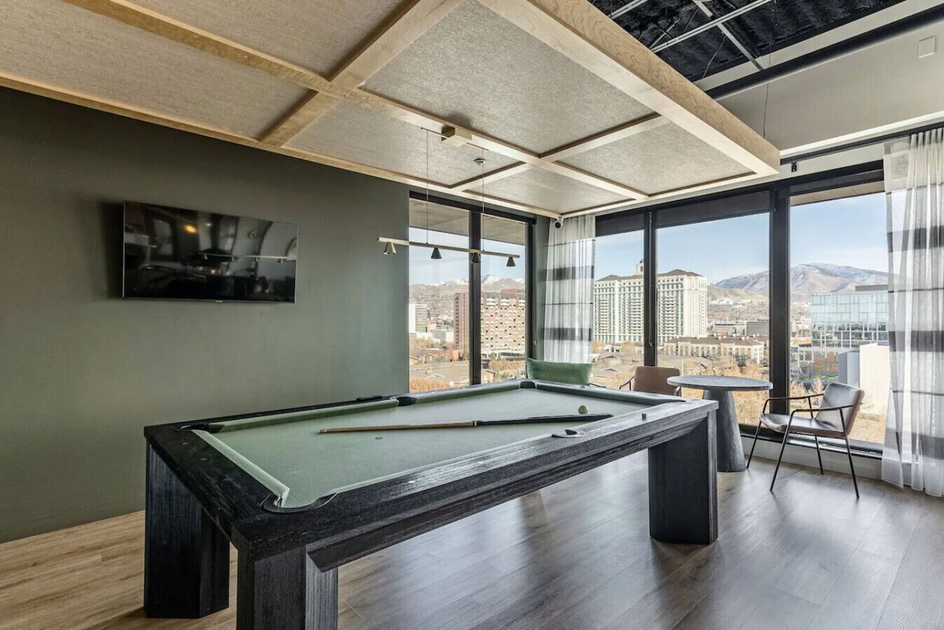 Una mesa de billar en una habitación con ventanas amplias, un televisor en la pared y vistas a un paisaje urbano.