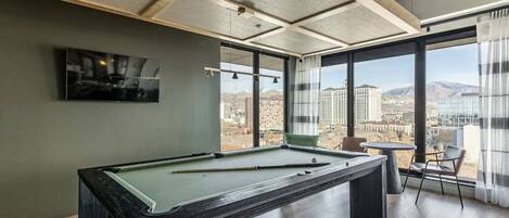 Una mesa de billar en una habitación con ventanas amplias, un televisor en la pared y vistas a un paisaje urbano.