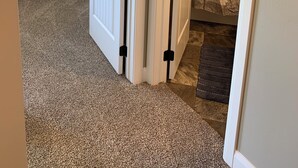A hallway with a carpeted floor, a door with black hardware, a small table with a lamp and a plant, and a patterned curtain in the background. - Luxury Townhouse in the Heart of Southern Utah (Cedar City)