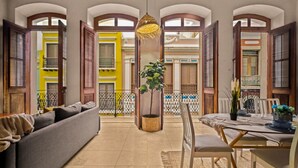 A spacious living area with a sofa, dining table, and chairs. There are open wooden shutters, a hanging light fixture, and a potted plant.