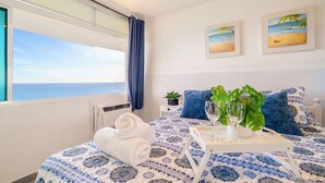 Un dormitorio con cama, ventana con vista al mar, dos cuadros en la pared y una mesita con planta y vasos.