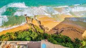 Vista aérea de una playa con aguas turquesas cristalinas, una orilla arenosa y abundante vegetación, incluyendo palmeras.