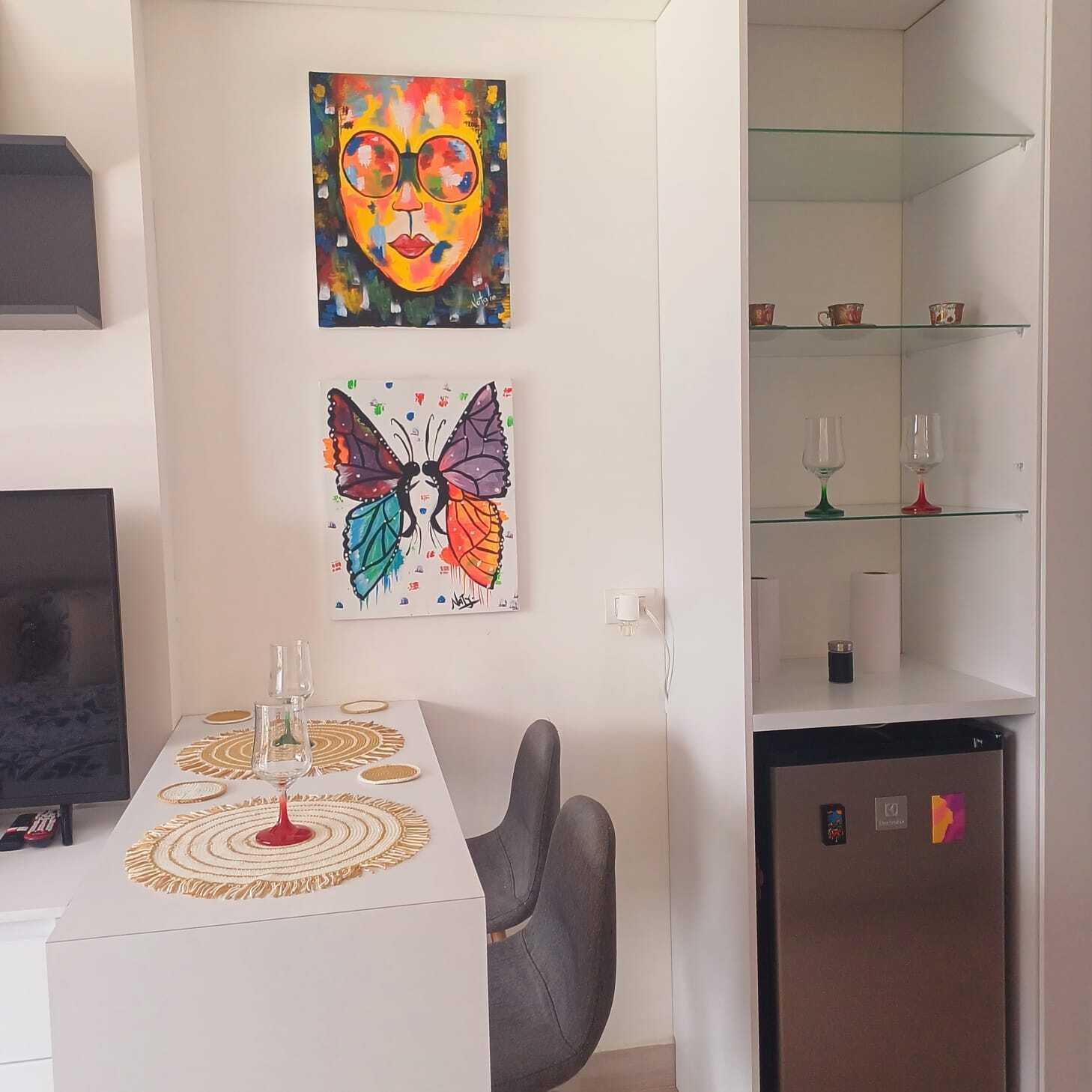 A modern living room with a dining table, two paintings, a glass cabinet, and a refrigerator.