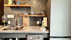 A kitchen with a marble countertop, a stainless steel sink, a wooden shelf, and a refrigerator.