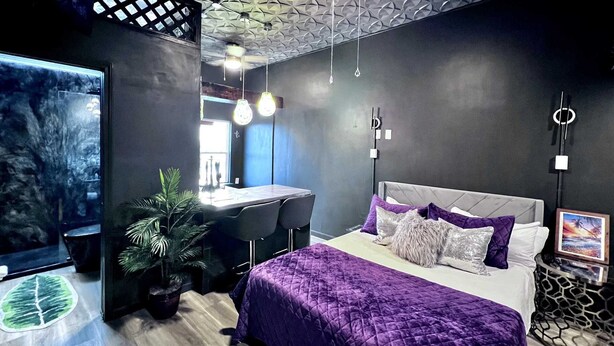 A modern bedroom with a large bed, a desk, and a chair. There is a potted plant and a painting on the wall.